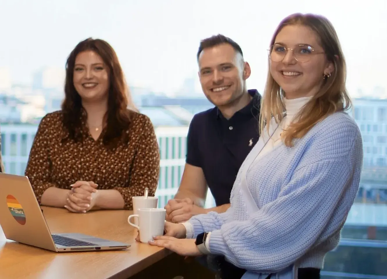 Drei lächelnde Mitarbeiter von 50Hertz Transmission GmbH in einem Büro bei einer Team-Besprechung mit Laptop und Kaffeetassen.