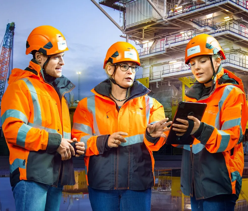 Three engineers in bright orange safety gear discussing plans on a tablet at an industrial site.