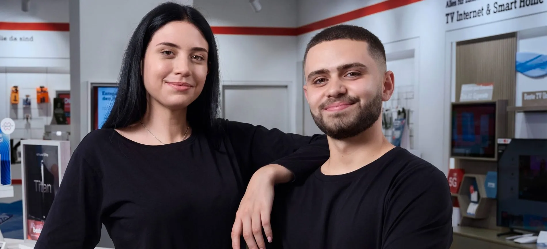 Zwei junge A1-Mitarbeitende lächeln im Telekommunikations-Shop in die Kamera. Du bist willkommen im Team.