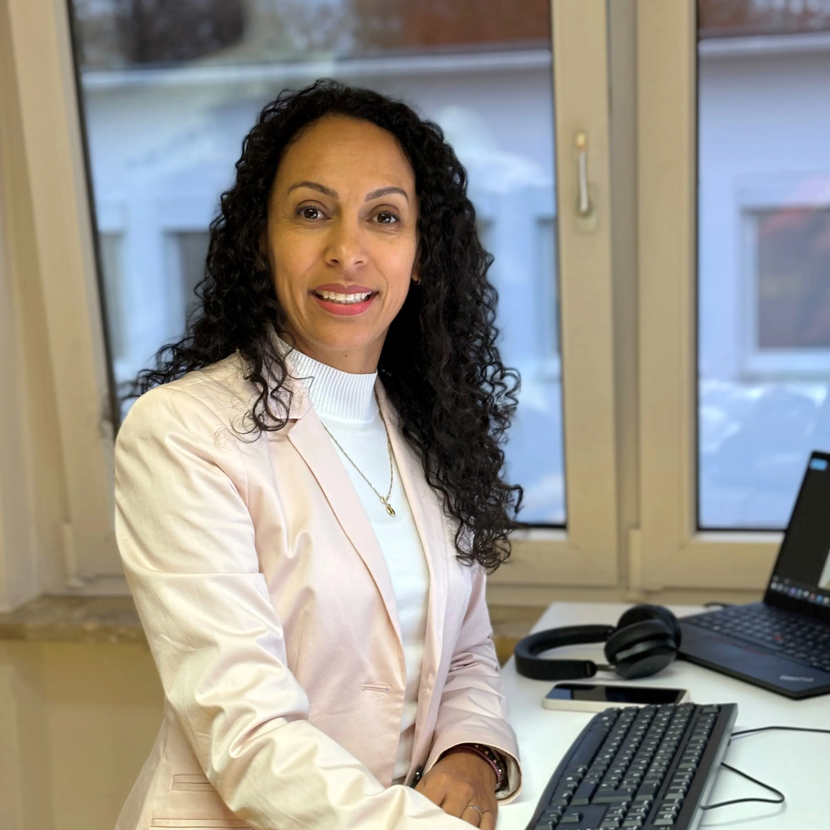Smiling woman in a light blazer at her desk with a laptop and headset, ready to welcome new team members at ABO Energy.