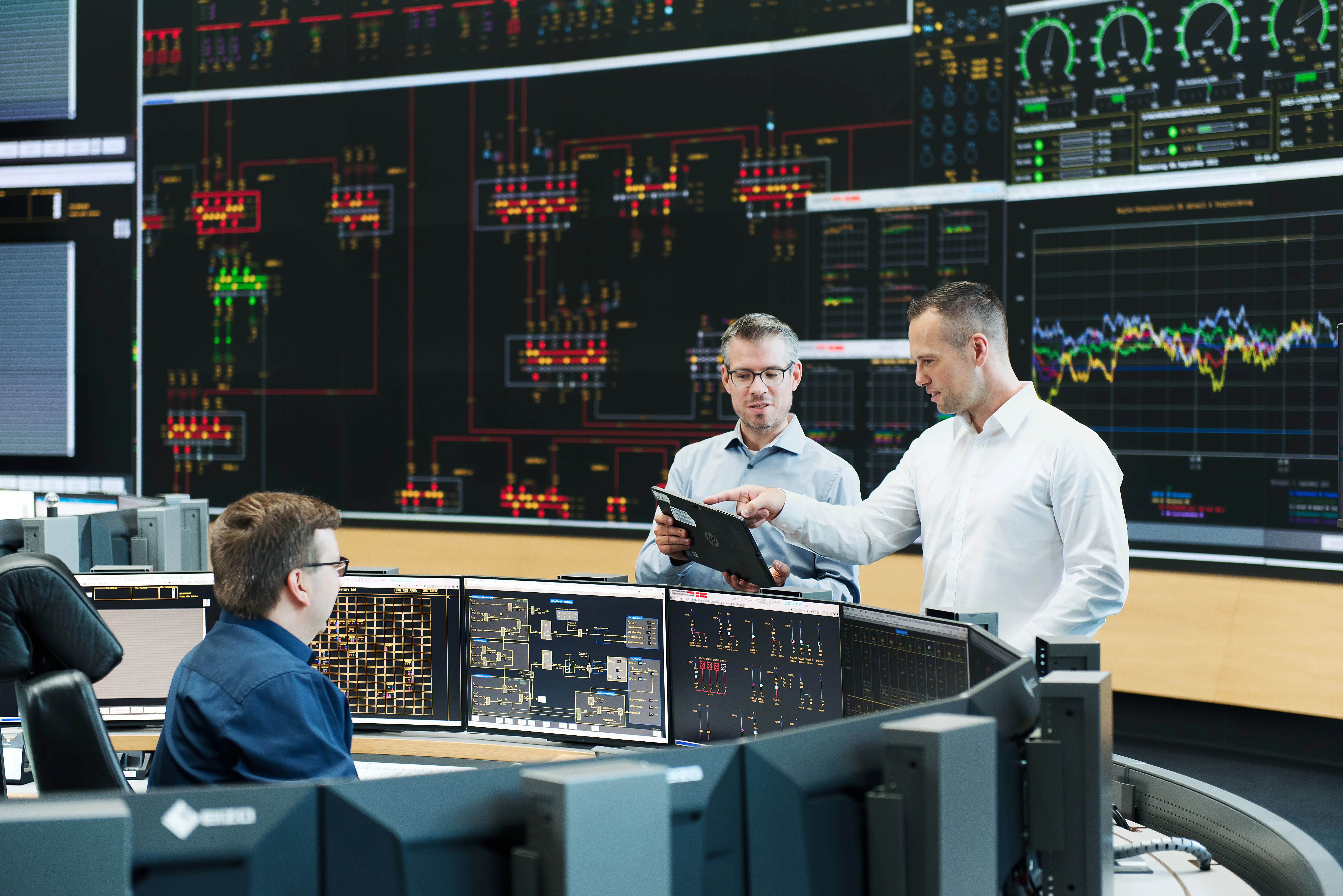 Ein Team von drei Personen arbeitet in einem modernen Kontrollraum von Amprion GmbH mit mehreren Bildschirmen und Energie-Diagrammen.