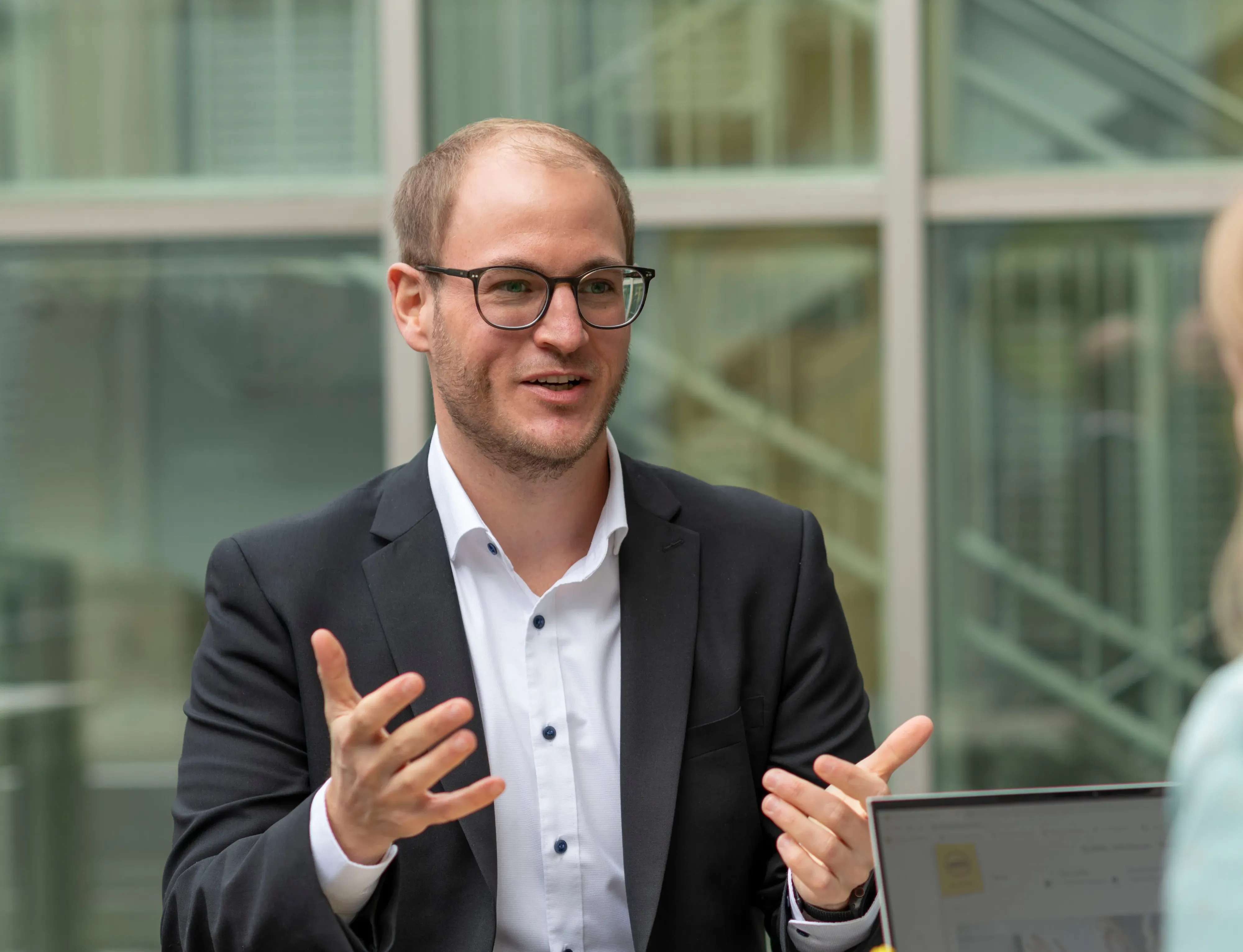 Ein Mann im Anzug spricht gestikulierend mit einer anderen Person, im Hintergrund ein modernes Bürogebäude.```