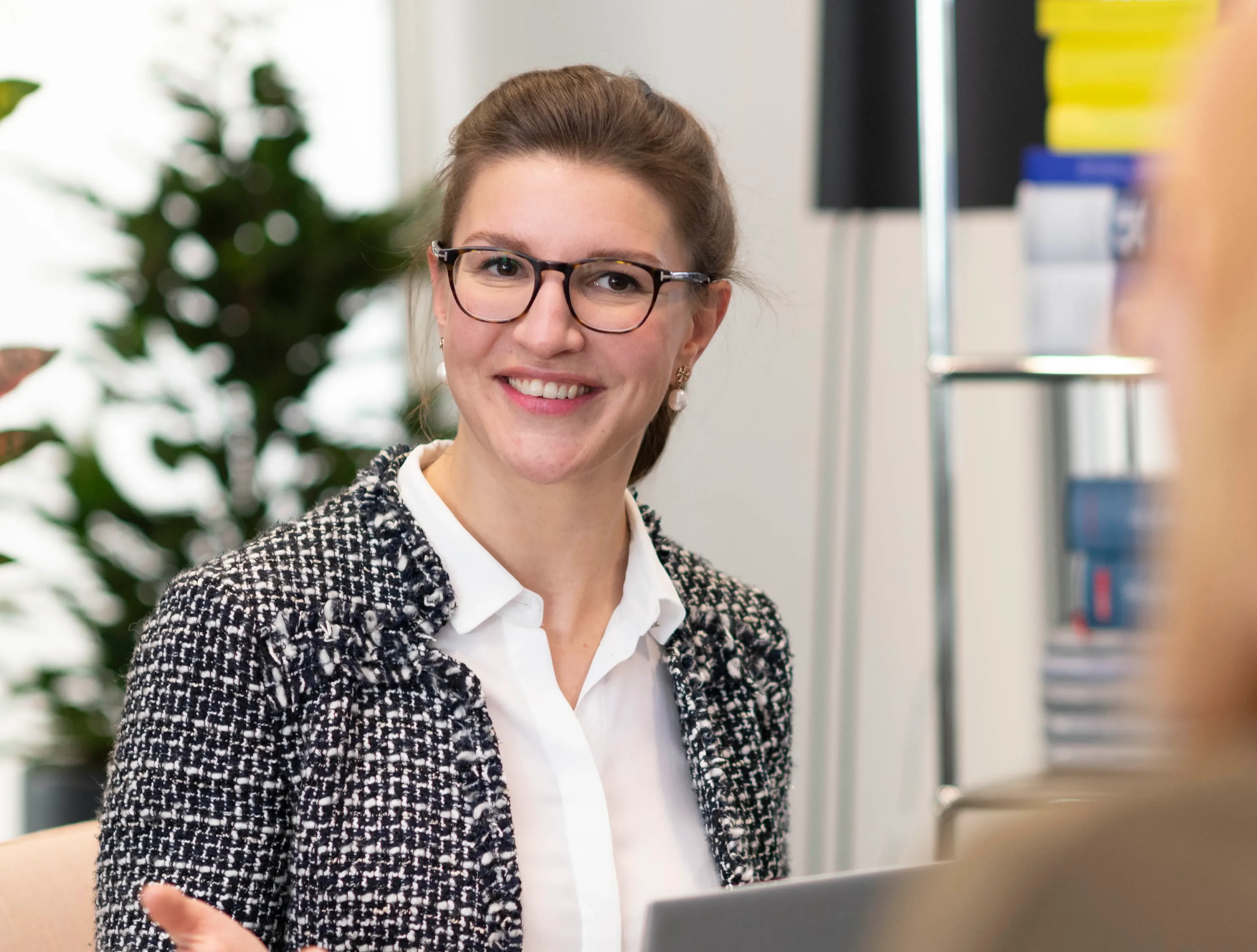 Eine lächelnde Frau in einem Büro spricht mit einer Kollegin. Im Hintergrund sind Pflanzen und Regale sichtbar.