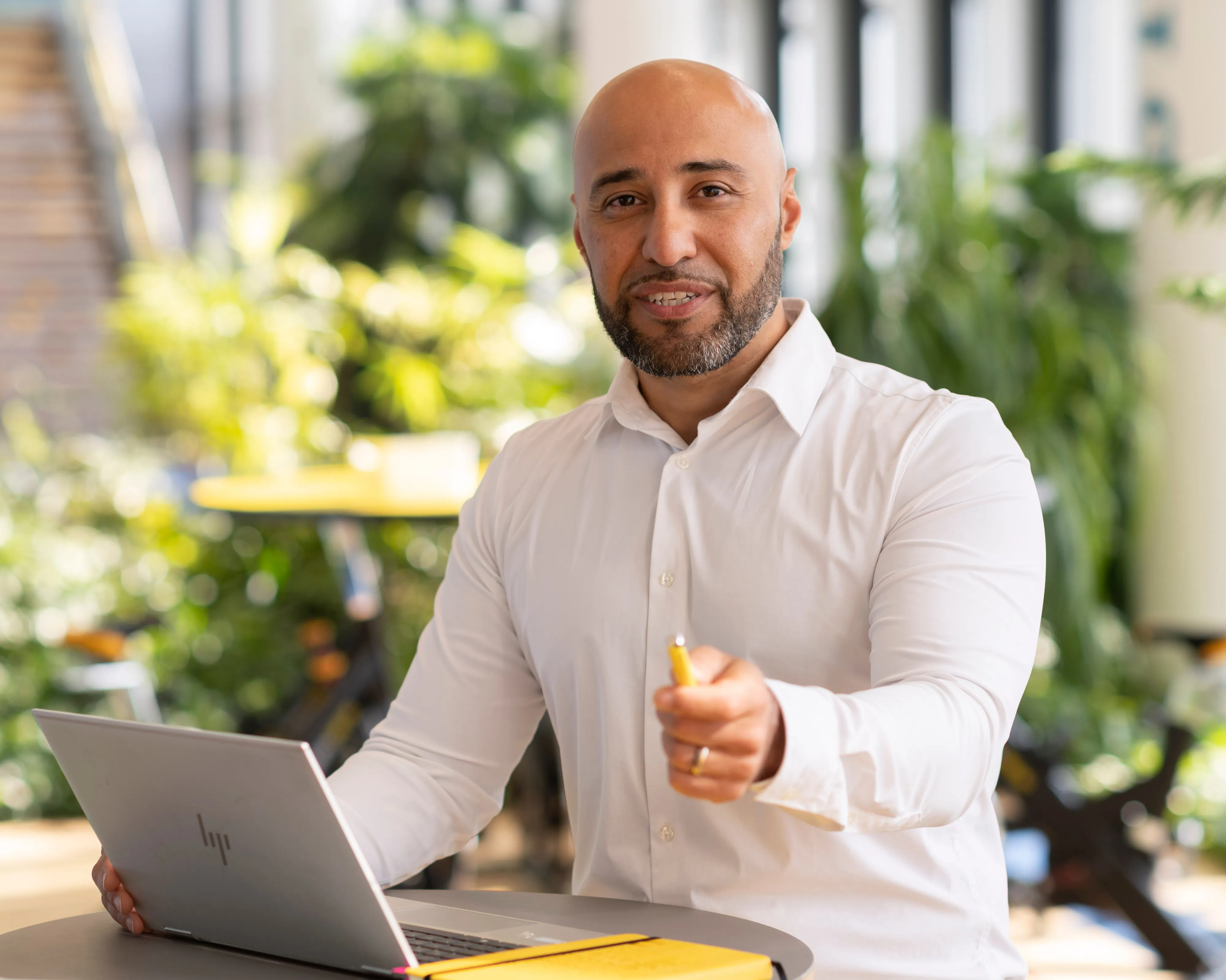 Ein Mann im weißen Hemd sitzt mit einem Laptop in einem modernen Büro und lächelt nachdenklich mit einem Stift in der Hand.