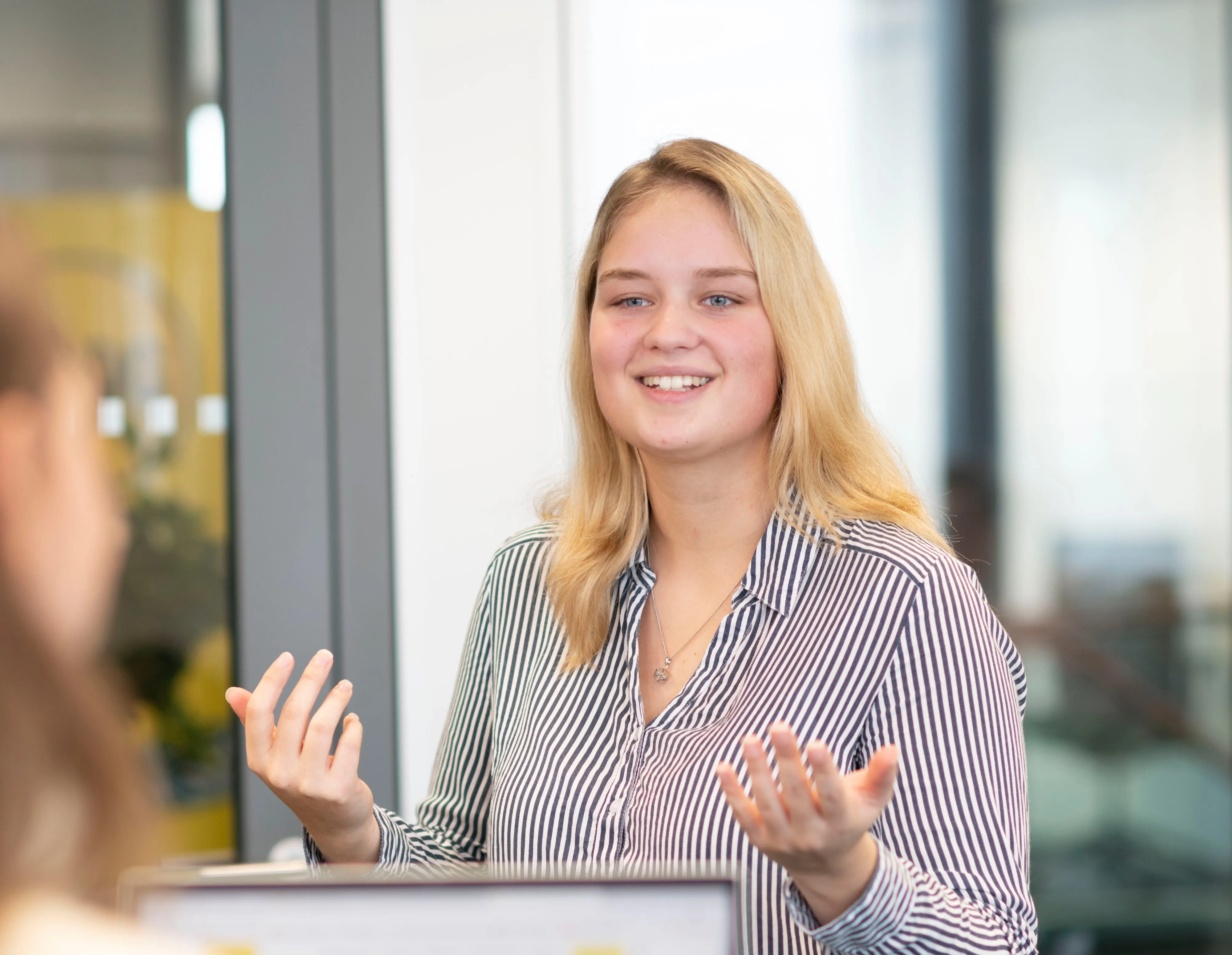 Eine lächelnde Frau in gestreiftem Hemd spricht mit Kollegen in einem modernen Büro.