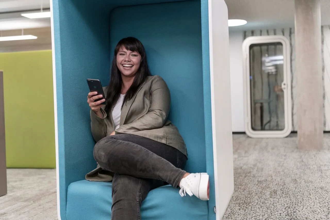 Eine lächelnde Frau sitzt entspannt in einer modernen Bürolounge und hält ein Smartphone in der Hand.