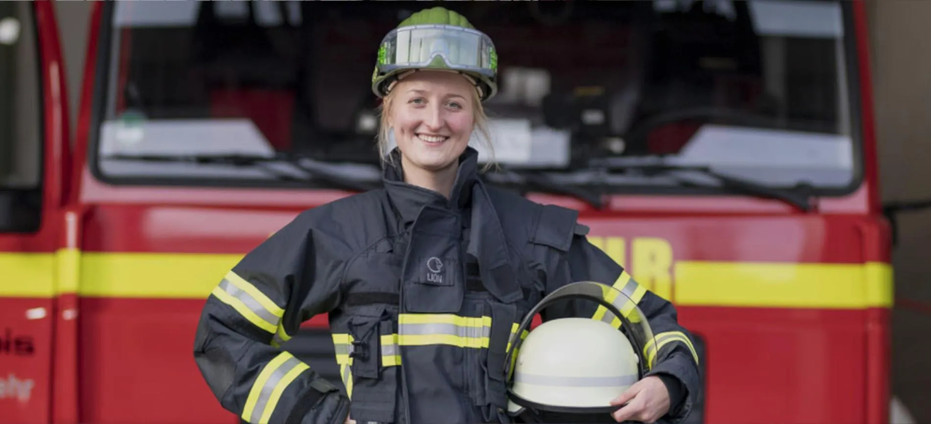 Junge Feuerwehrfrau in Einsatzkleidung steht lächelnd vor einem Feuerwehrfahrzeug und hält ihren Helm in der Hand.