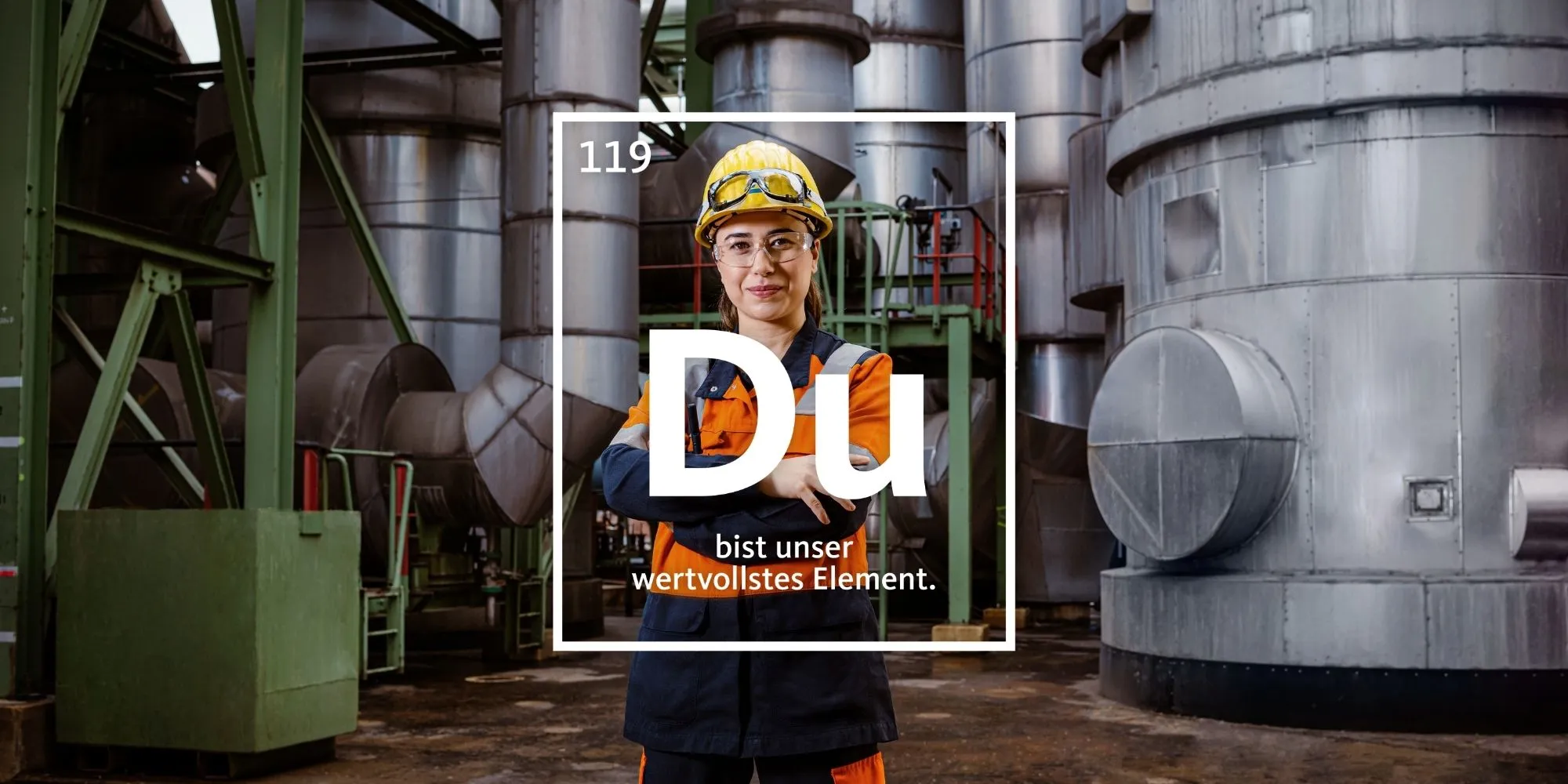 Ein Mitarbeiter mit Schutzkleidung steht lächelnd in einer Produktionshalle von Aurubis mit dem Slogan „Du bist unser wertvollstes Element“.