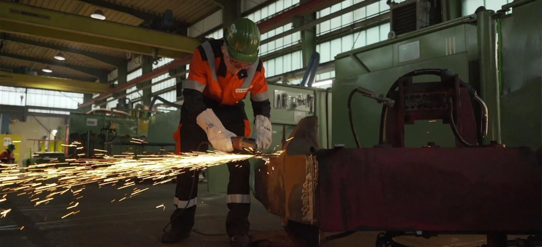 Du siehst einen Mitarbeitenden bei Aurubis AG, der mit Schutzkleidung Metall in einer Werkshalle schleift und Funken sprühen lässt.