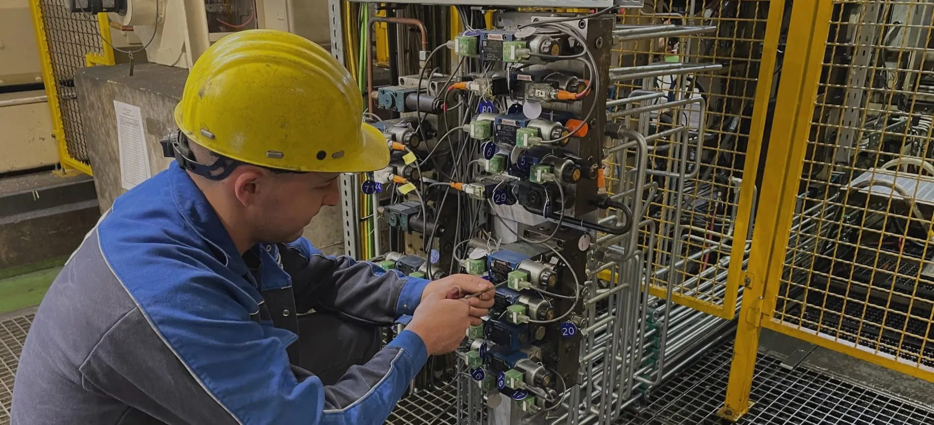 Du siehst einen Techniker mit Schutzhelm, der bei Aurubis eine industrielle Anlage wartet und elektrische Komponenten prüft.
