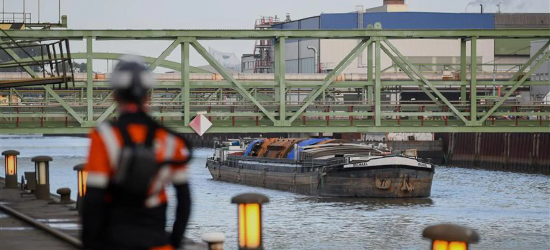 Du blickst auf ein Frachtschiff am Werkgelände von Aurubis, während eine Person in Schutzkleidung am Ufer steht.