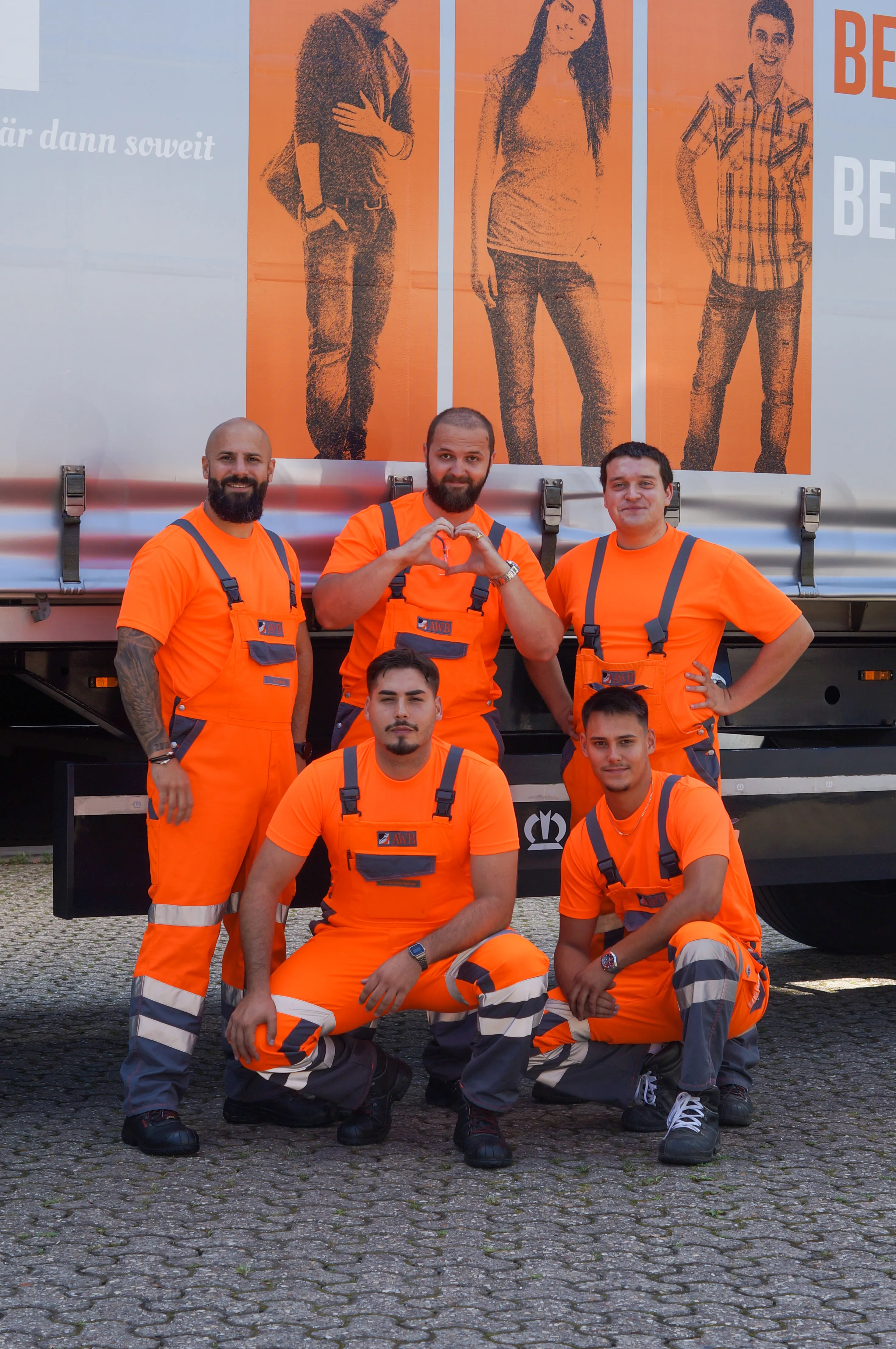 Fünf Kollegen in AWB-Arbeitskleidung posieren freundlich vor einem Müllfahrzeug und laden Dich ins Team ein.