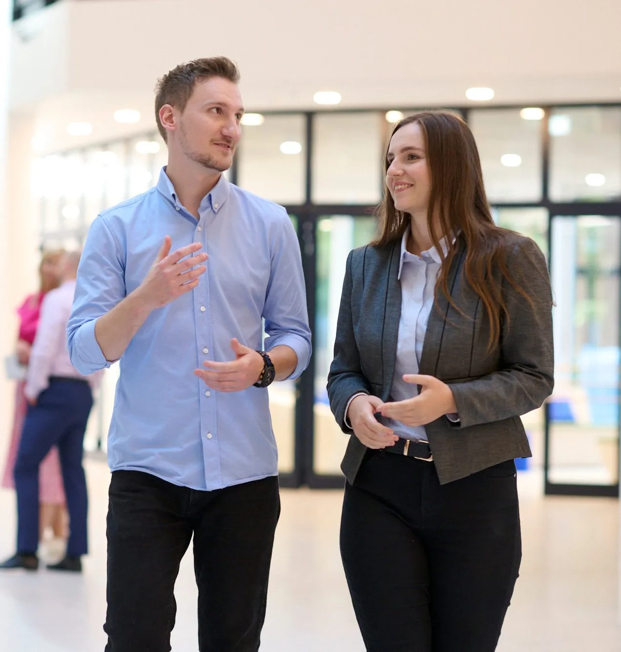 Eine lächelnde Frau in einem Bürogebäude mit Blick auf die Stadtlandschaft im Hintergrund.