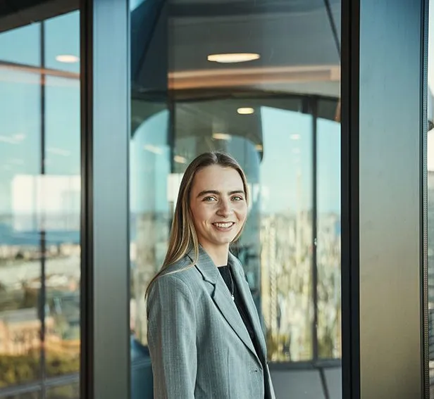 Eine lächelnde Frau in einem Bürogebäude mit Blick auf die Stadtlandschaft im Hintergrund.