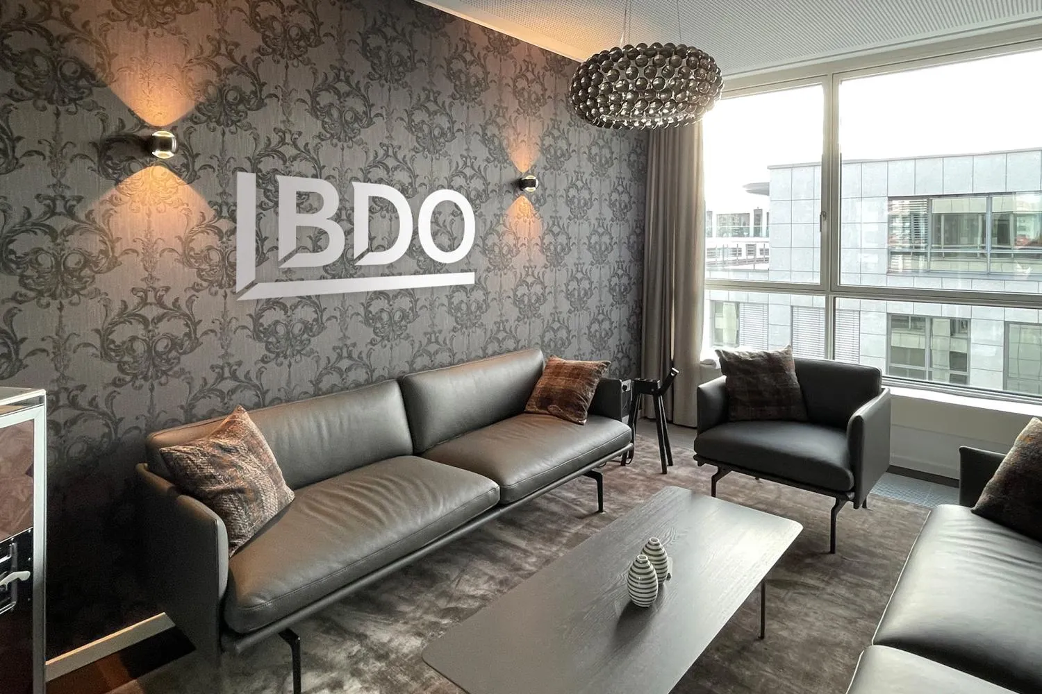 Moderner Lounge-Bereich bei BDO mit Ledersofas, großem Fenster und stilvoller Wand, ideal für Deine Pausen im Büro.