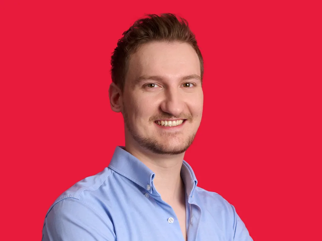 Junger Mann im blauen Hemd lächelt freundlich vor rotem Hintergrund, Karriere bei BDO im Bereich Wirtschaftsprüfung.