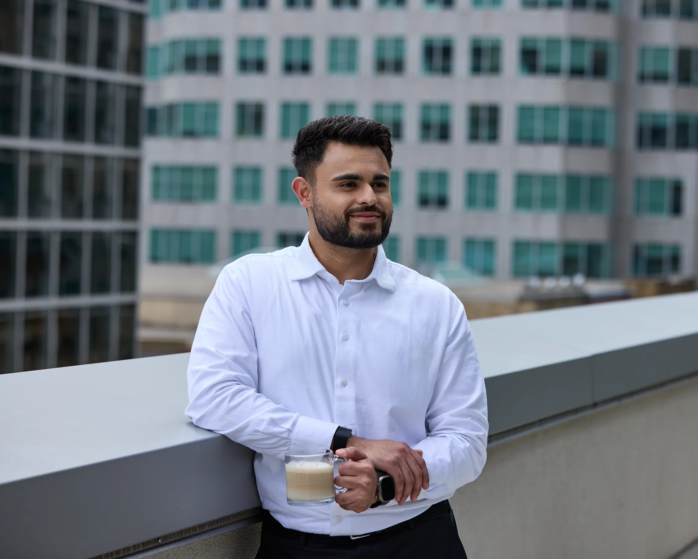 Junger Mann in weißem Hemd steht mit Kaffee auf einer Büro-Terrasse und blickt entspannt in die Ferne.