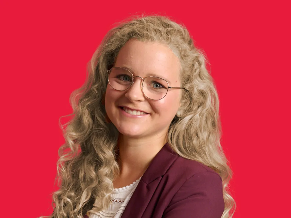 Junge Frau mit langen lockigen Haaren und Brille lächelt freundlich vor rotem Hintergrund, passend zur Karriere bei BDO.