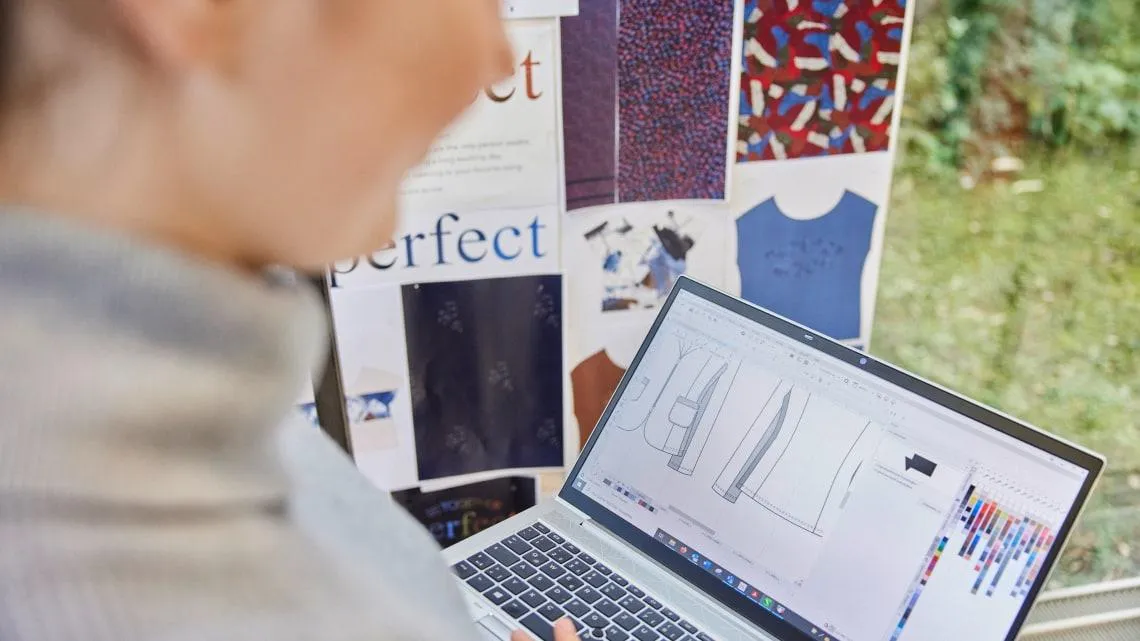 Du siehst eine Person, die am Laptop Modedesigns erstellt, mit Stoffmustern und Skizzen im Hintergrund.