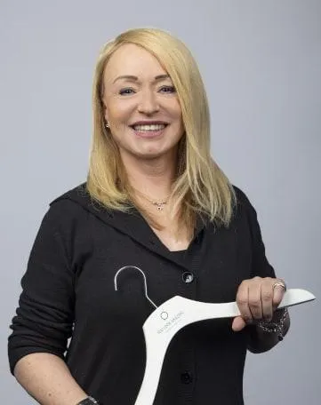 Lächelnde Frau im schwarzen Outfit hält einen Kleiderbügel mit Betty Barclay Logo – Arbeiten im Einzelhandel.