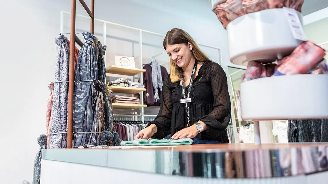Mitarbeiterin faltet Kleidung und lächelt im Betty Barclay Store – werde Teil unseres Retail-Teams!