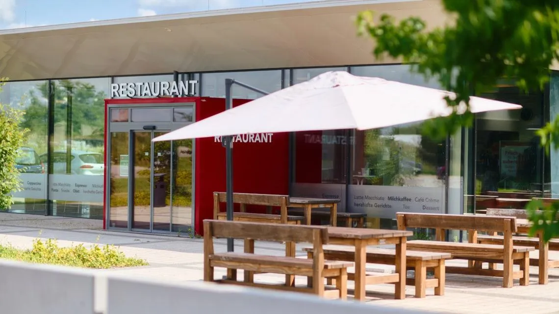 Außenbereich eines modernen Restaurants mit Holzsitzbänken und Sonnenschirm, ideal für Deine Mittagspause bei Betty Barclay.