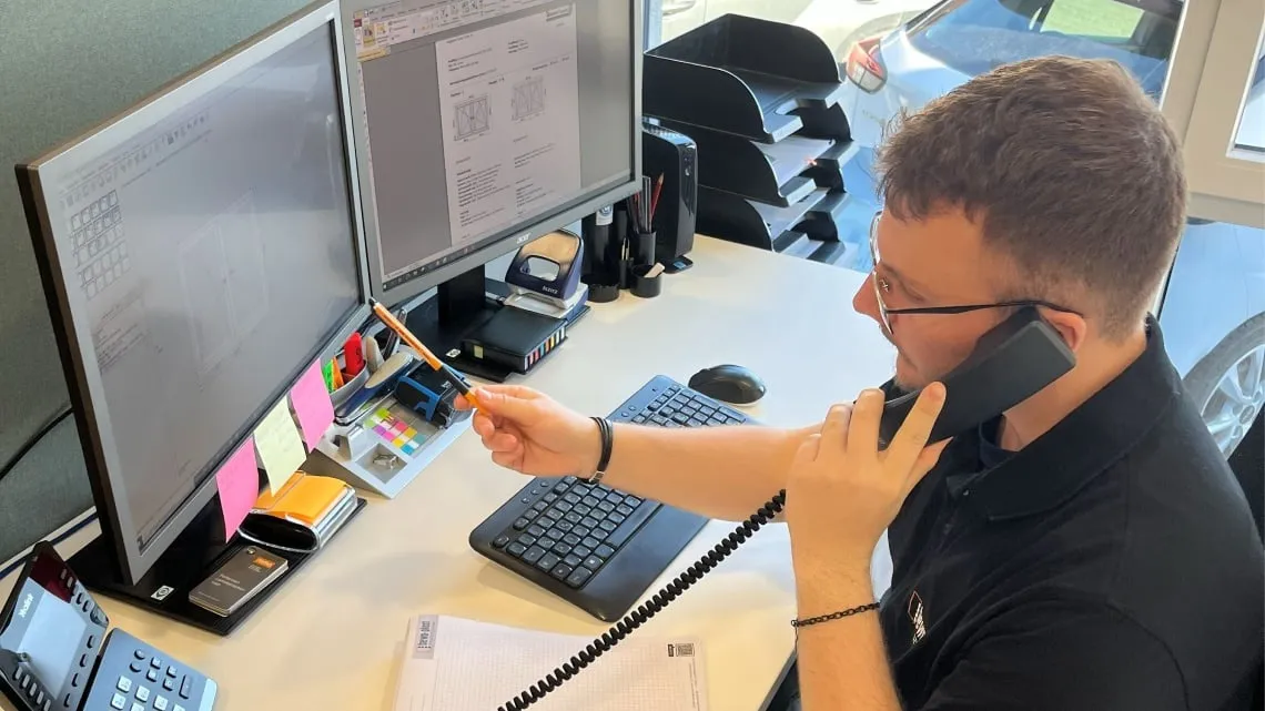 Du siehst einen Mitarbeitenden, der am Schreibtisch am Telefon ist und dabei auf technische Zeichnungen am Computer zeigt.