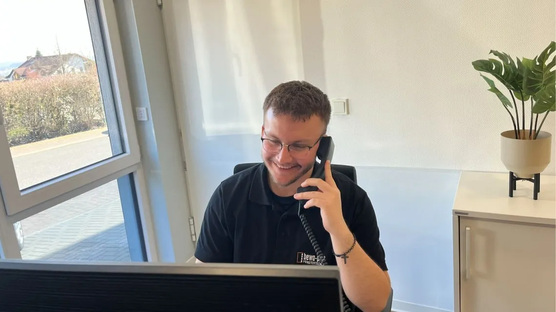 Junger Mitarbeiter von bewa-plast Beck GmbH telefoniert lächelnd am Schreibtisch im modernen Büro.