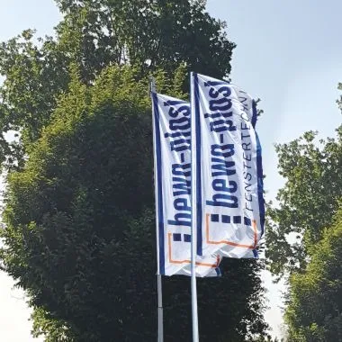 Zwei Fahnen mit dem Logo von bewa-plast Beck GmbH Fenstertechnik wehen vor Bäumen im Außenbereich.