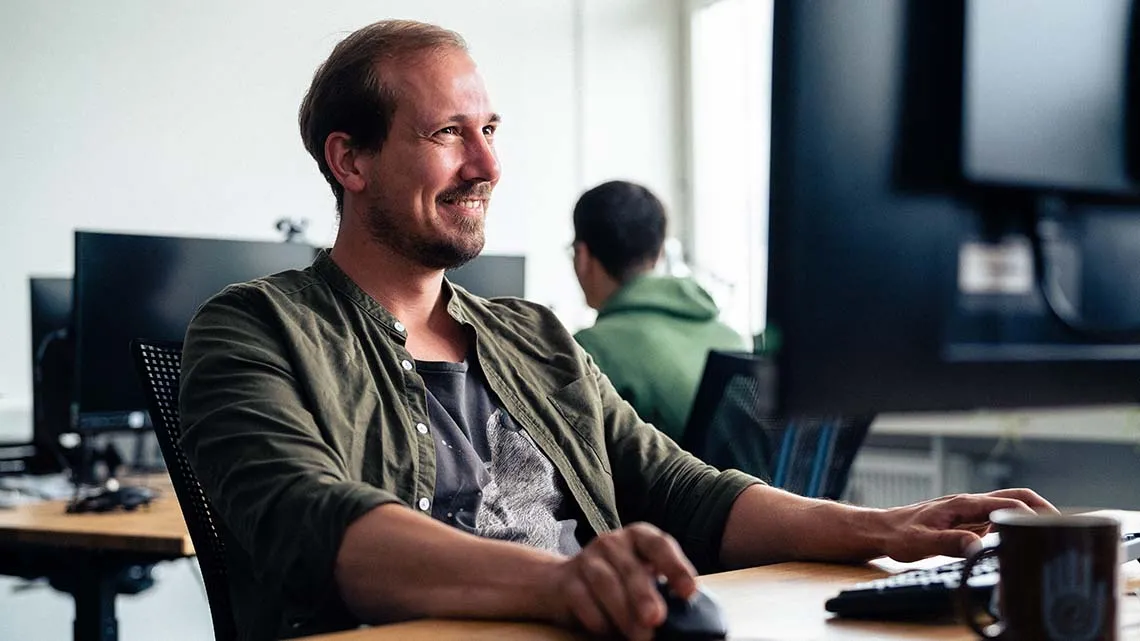 Mitarbeiter arbeitet lächelnd am Computer im modernen Büro bei BIKE24, freundliche Arbeitsatmosphäre im Retail-Bereich.