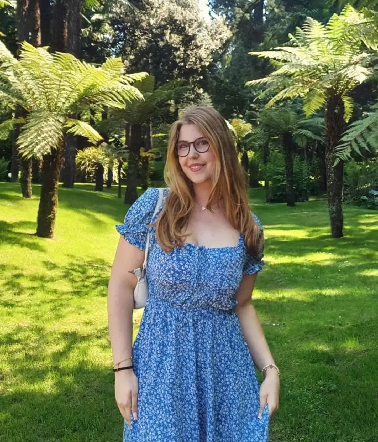 Junge Frau mit Brille und blauem Kleid lächelt in einem sonnigen Park, umgeben von grünen Palmen und Farnen.