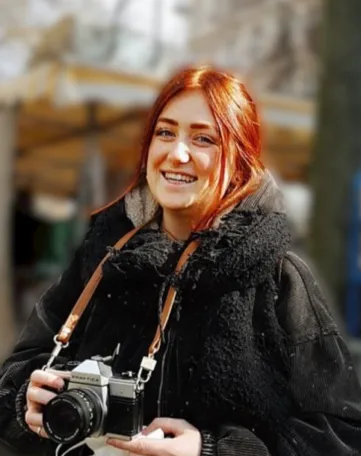 Junge Frau mit rotem Haar lächelt in die Kamera und hält einen Fotoapparat, Symbol für kreative Karrieremöglichkeiten bei mkk.
