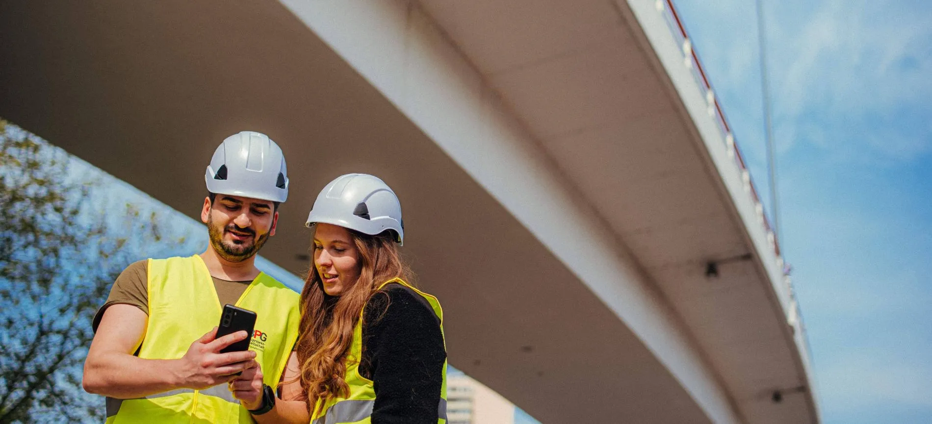 Zwei Bauingenieure mit Schutzhelmen und Warnwesten schauen gemeinsam auf ein Smartphone unter einer Brücke.