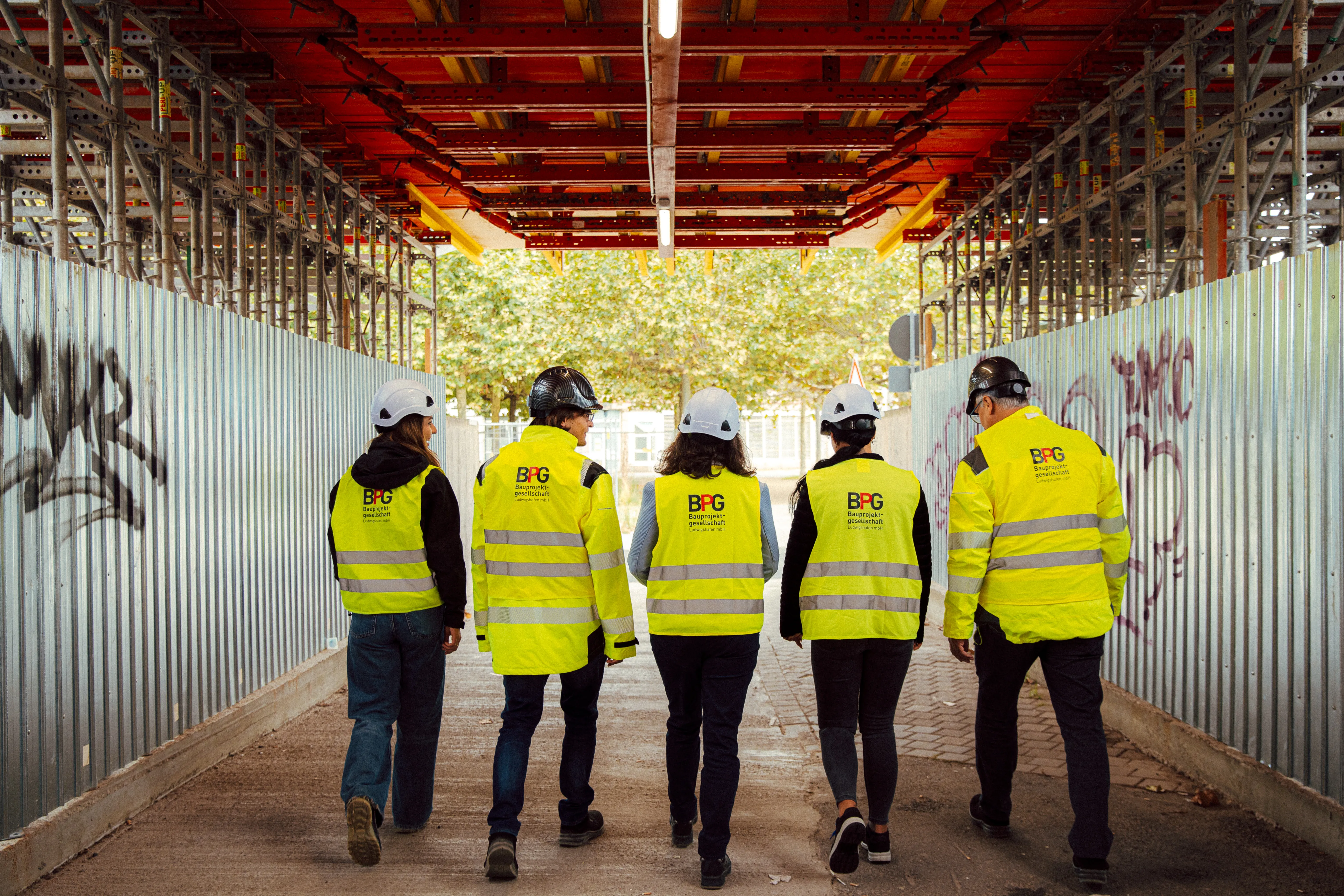 Fünf BPG-Mitarbeitende mit Schutzhelmen und Warnwesten gehen gemeinsam über eine Baustelle.