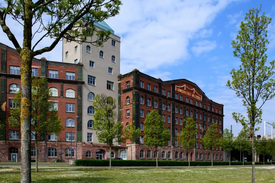Backstein-Gebäude der Ludwigshafener Holzwerke mit begrüntem Vorplatz und blauem Himmel – hier kannst Du mitgestalten.