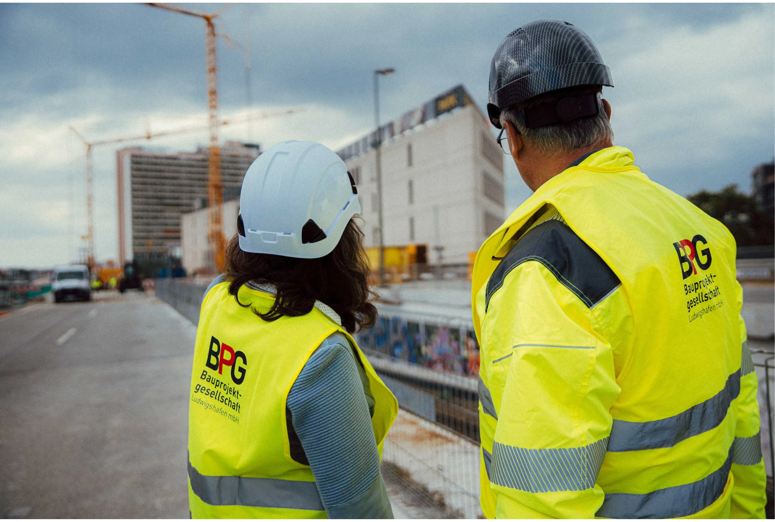 Zwei Bauleiter:innen der BPG Ludwigshafen schauen auf eine Baustelle mit Kränen und Rohbau im Hintergrund.