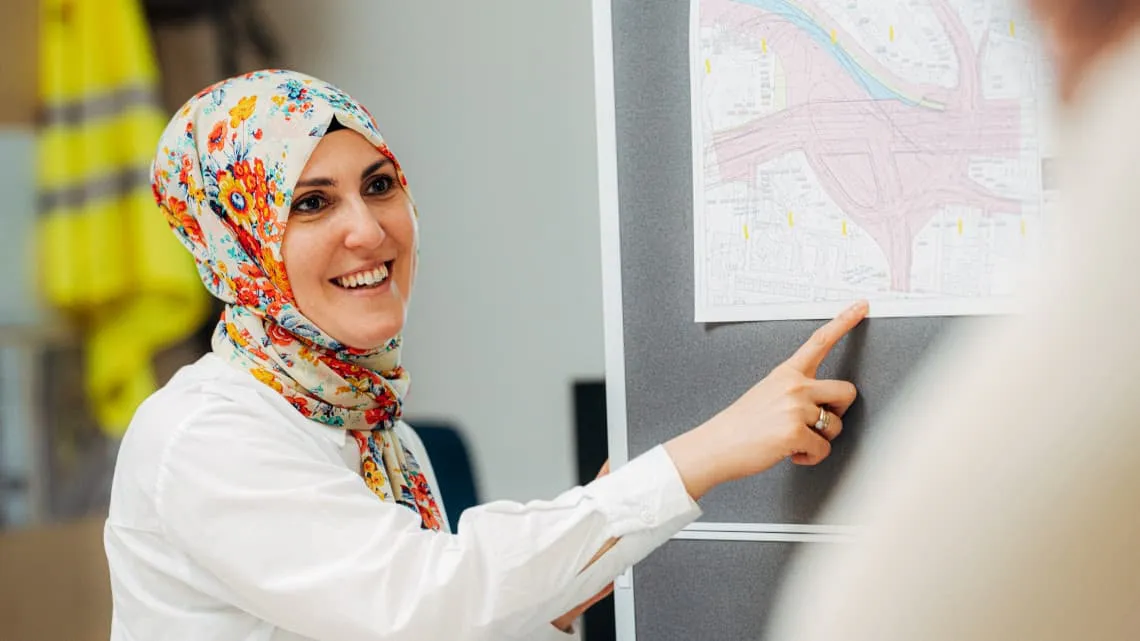 Junge Frau zeigt lächelnd auf einen Bauplan und erklärt Dir ein Bauprojekt im Team bei Bauprojektgesellschaft Ludwigshafen.