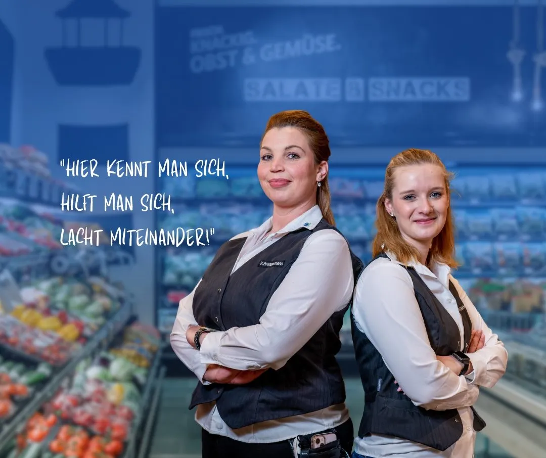 Zwei Mitarbeiterinnen von Combi lächeln in einem Supermarkt vor der Gemüseabteilung.