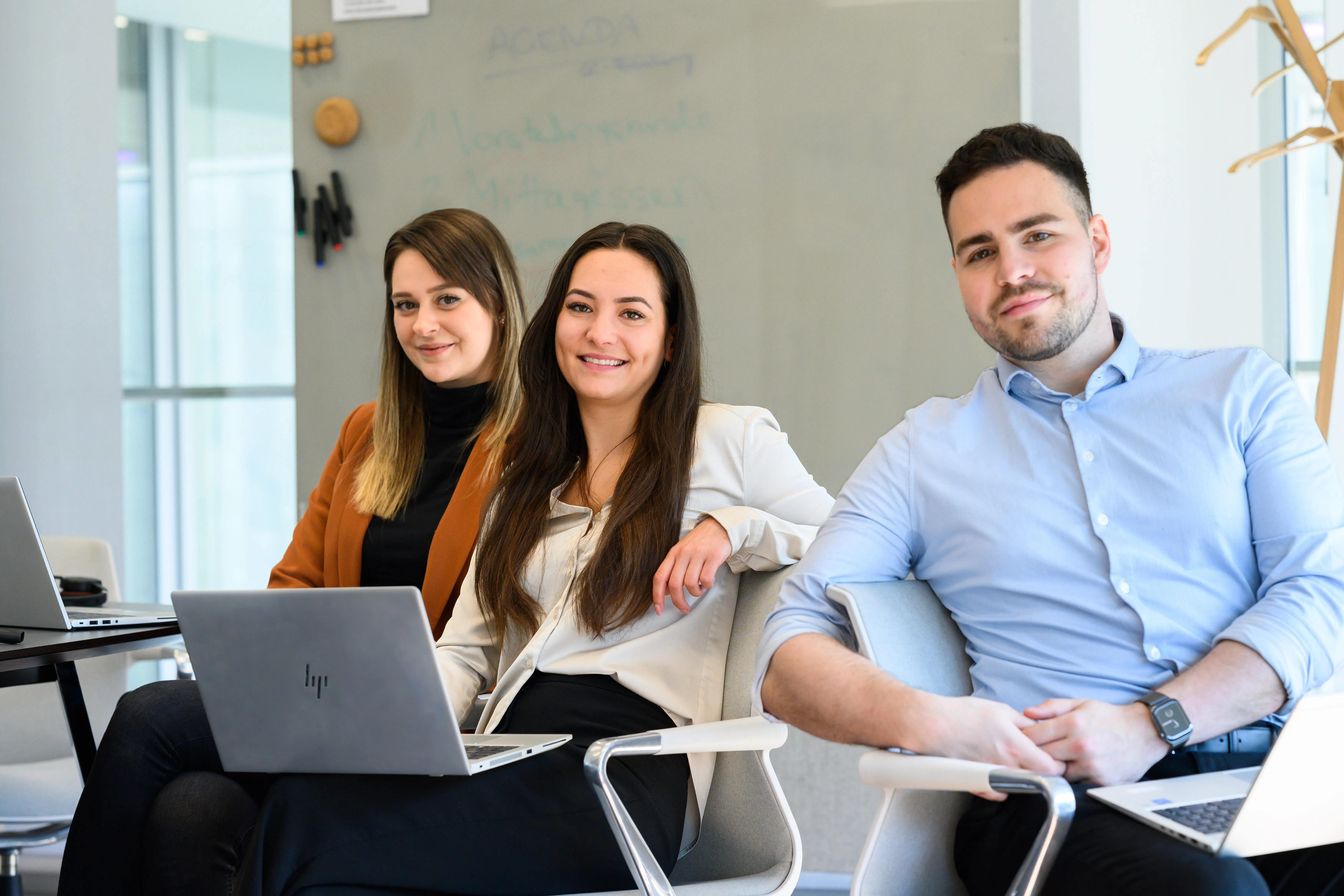 Three young professionals smiling, sitting with laptops in a modern office at Computacenter AG & Co. oHG.