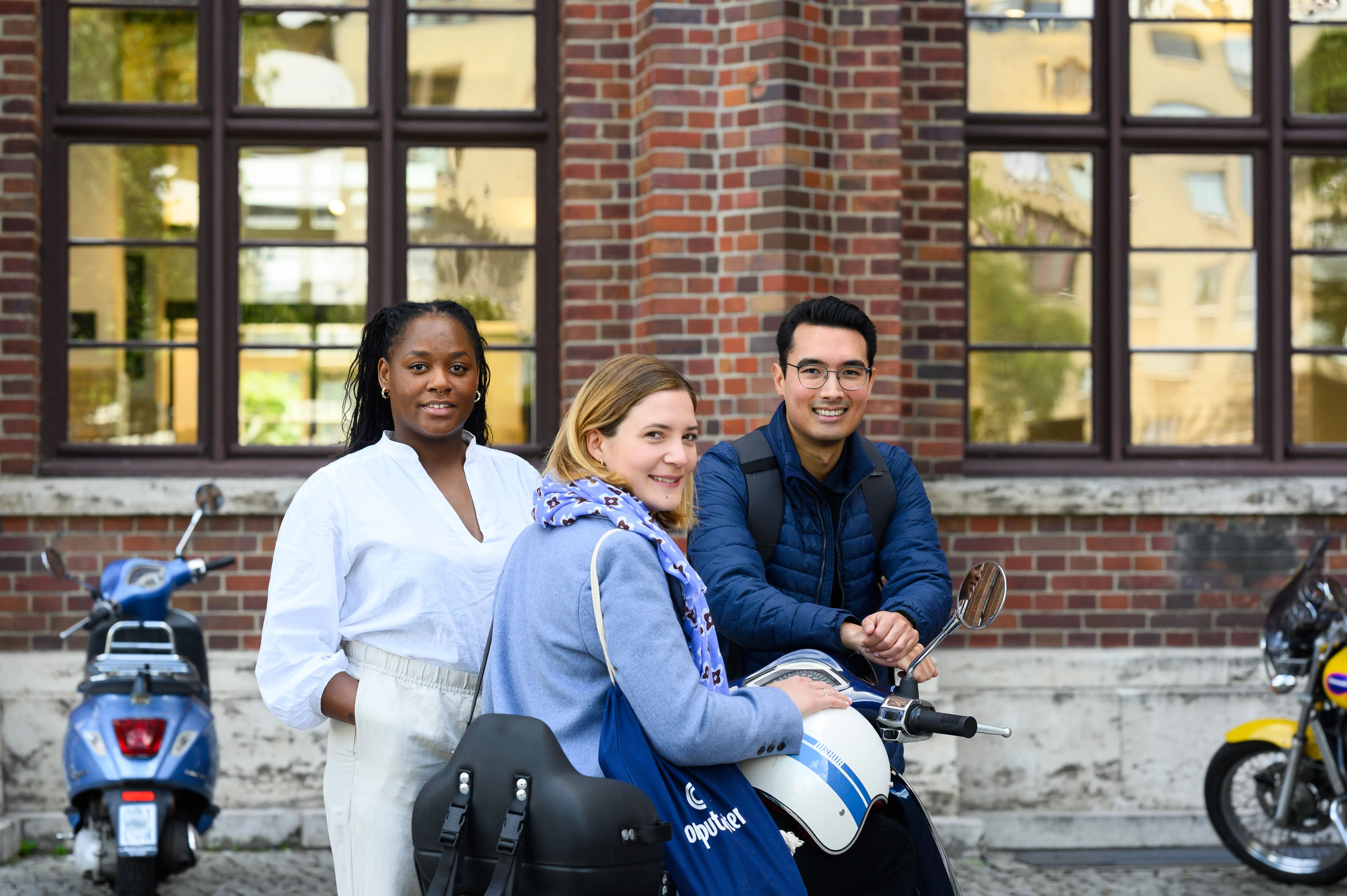Alt Texa woman with long brown hair, wearing a white blouse, sitting in an office environment. She has a pleasant smile and appears to be in front of computer monitors, with the office space in the background blurred out.t