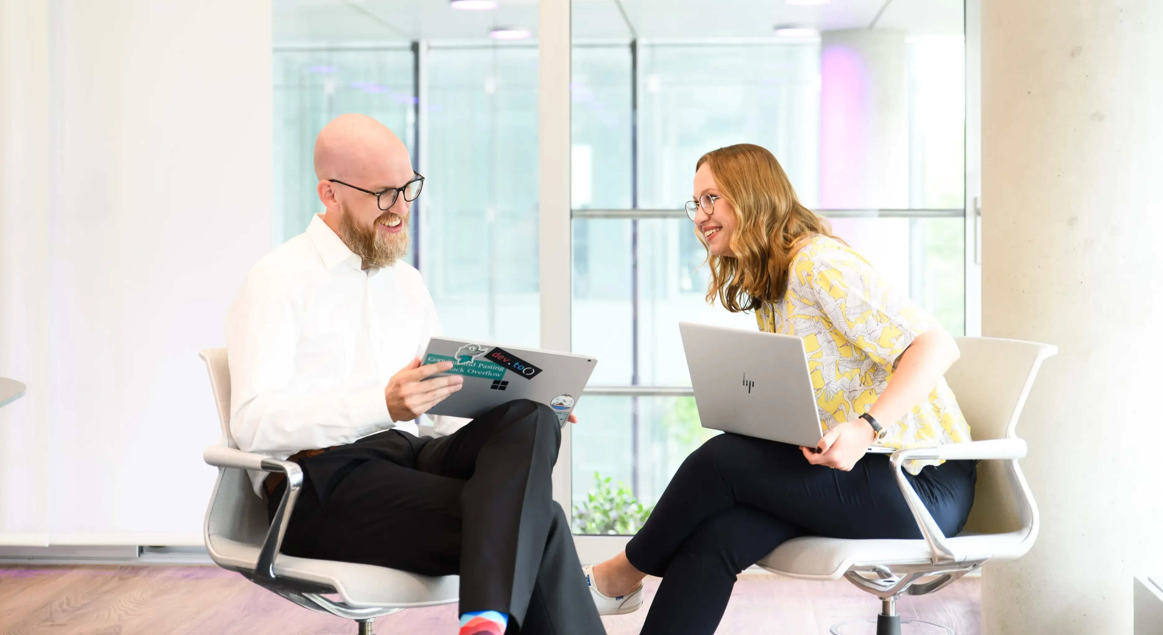 Zwei IT-Profis sitzen in einem modernen Büro und diskutieren lächelnd über Laptops.