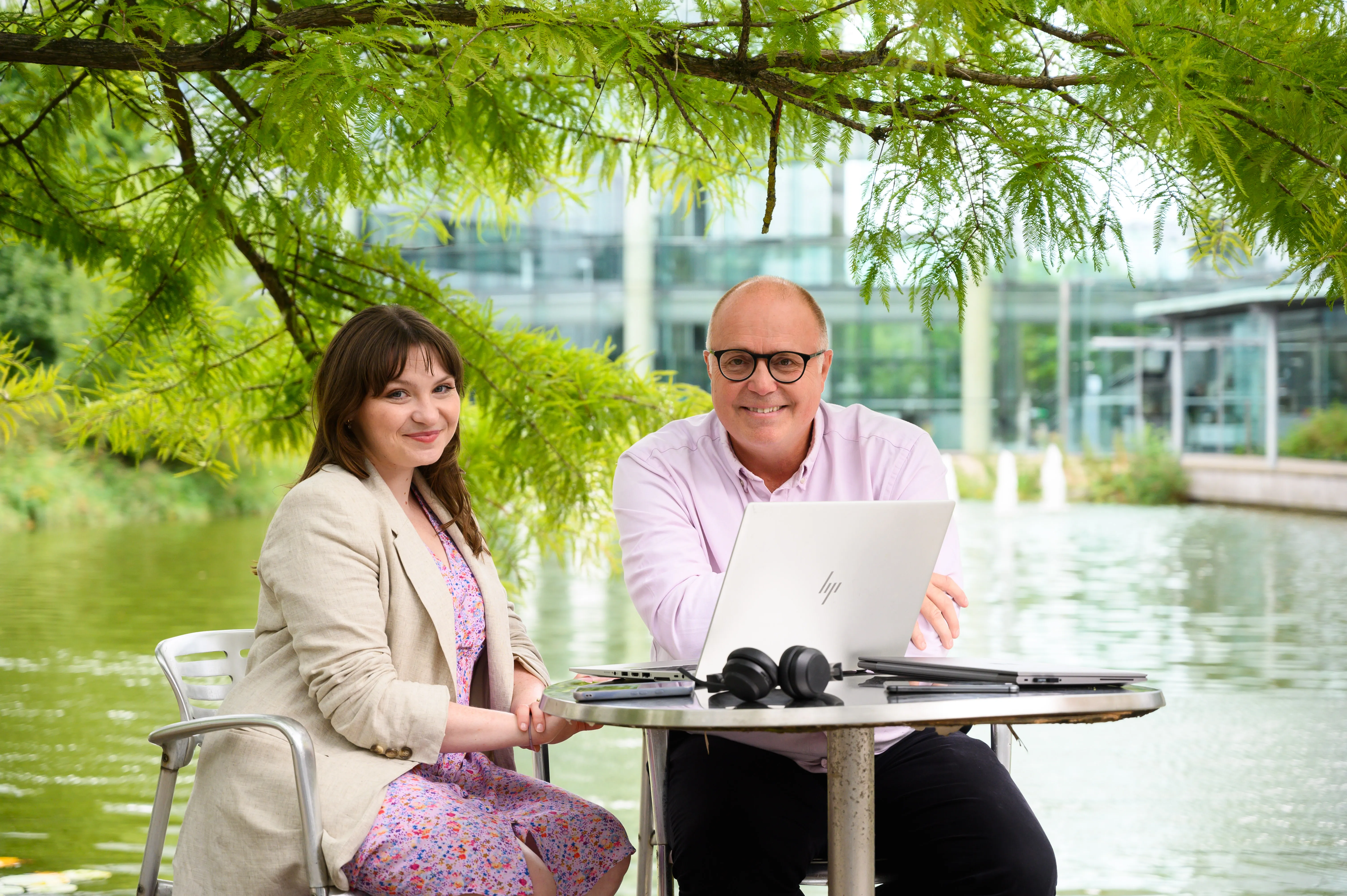 Two Computacenter AG employees enjoying an outdoor workspace by a pond, collaborating on a laptop under a tree.