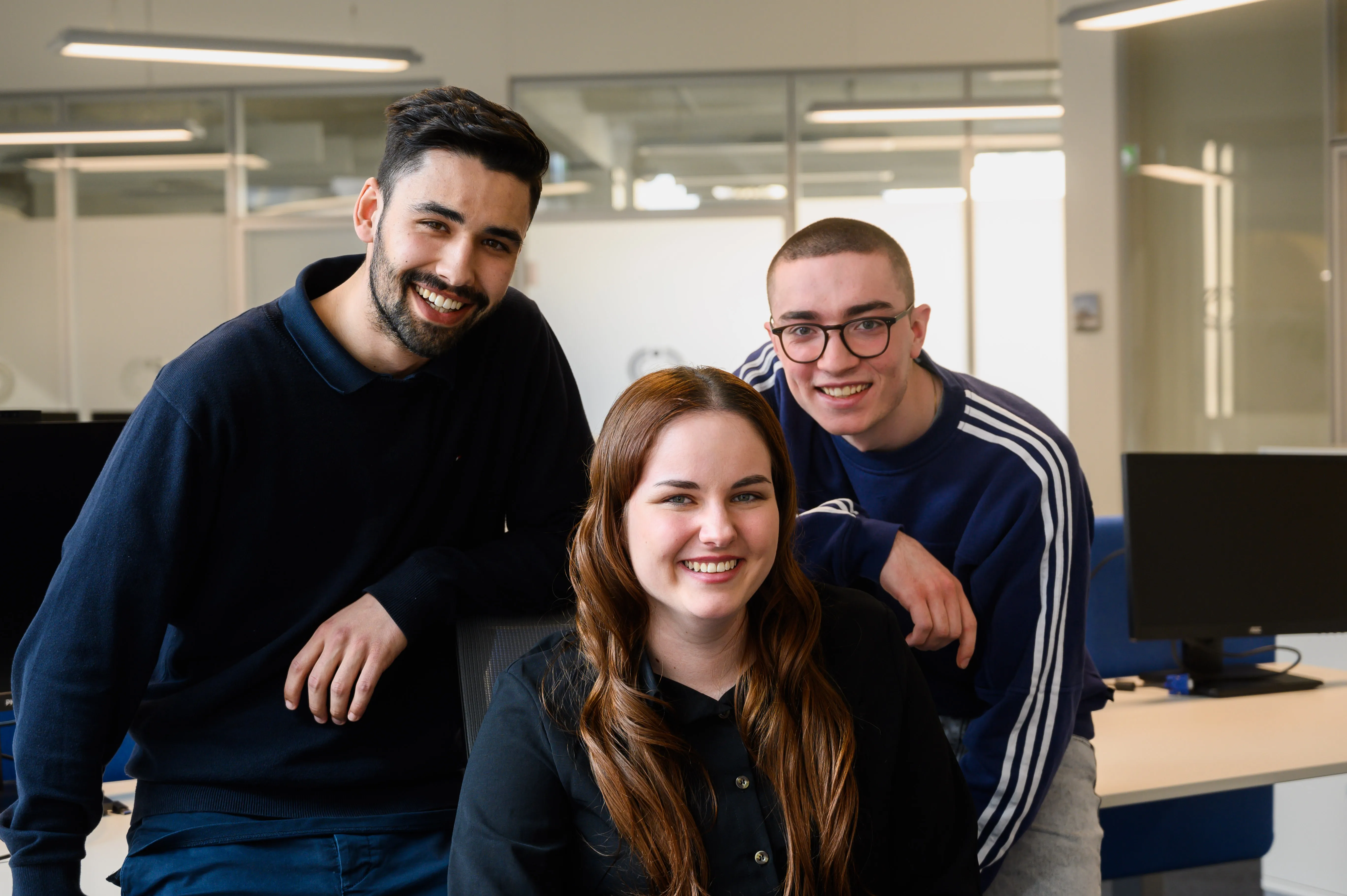 Alt Texa woman with long brown hair, wearing a white blouse, sitting in an office environment. She has a pleasant smile and appears to be in front of computer monitors, with the office space in the background blurred out.t