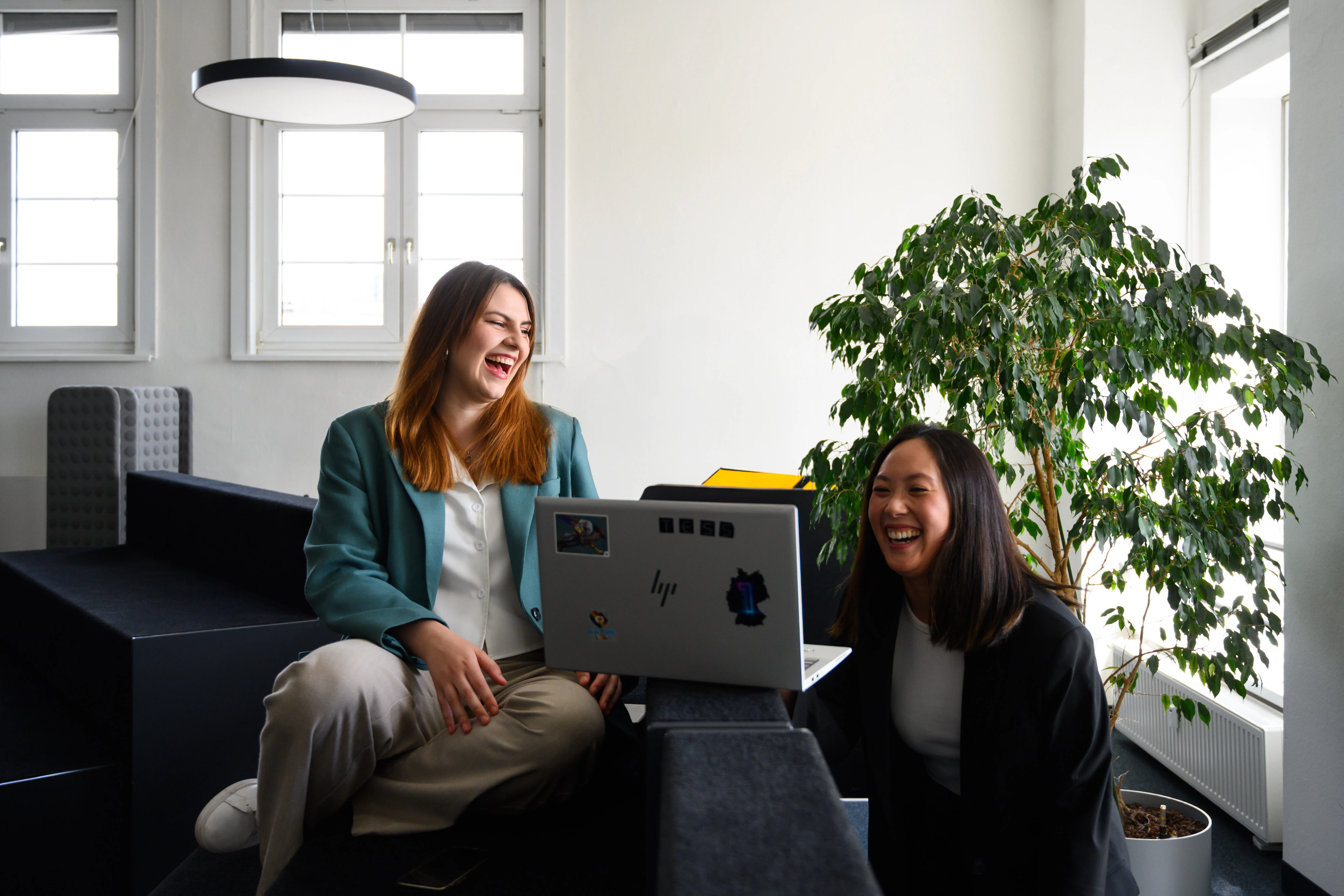 Zwei lachende Frauen arbeiten gemeinsam an einem Laptop in einem modernen Büro.
