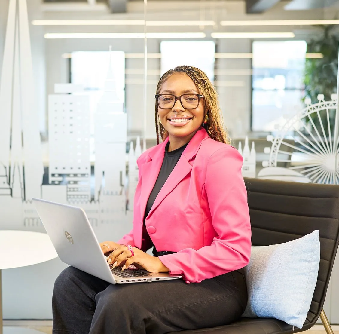 Smiling professional in a bright pink blazer working on a laptop in a modern, tech-savvy office.