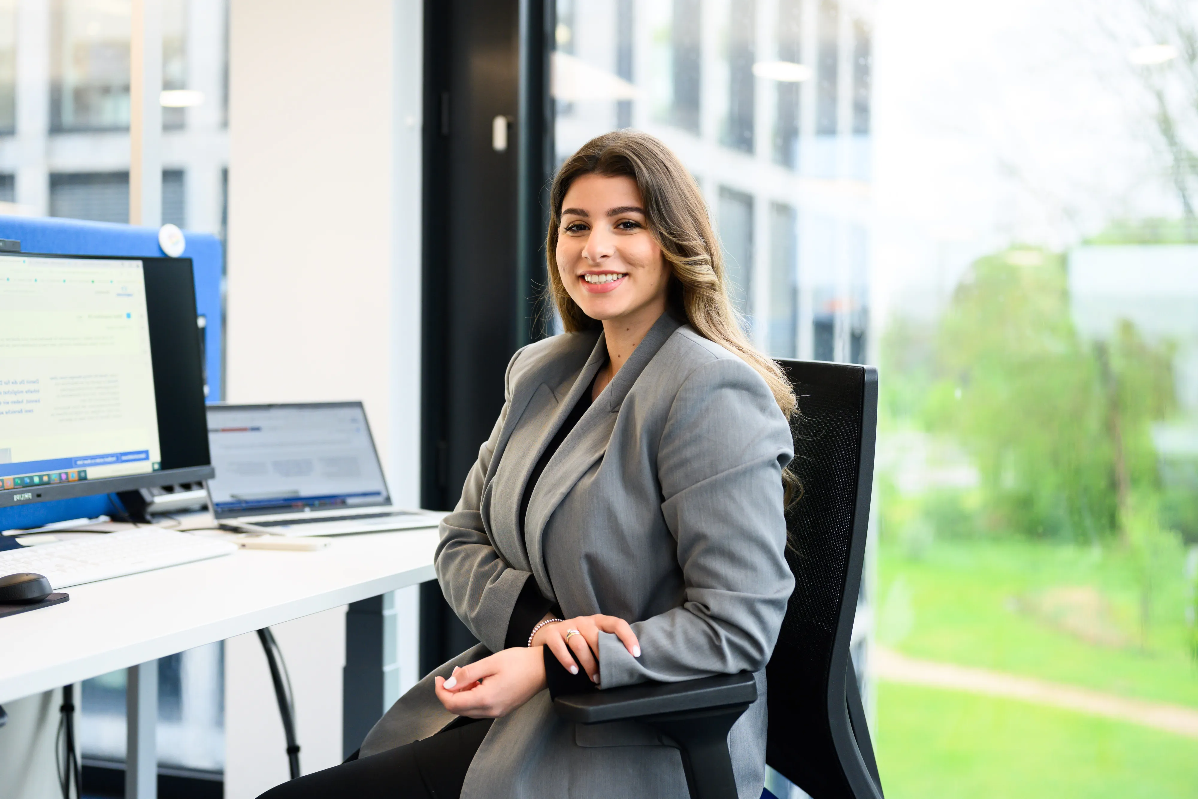 A man wearing glasses, a light purple shirt, and dark blue pants sits on a wicker chair outdoors, smiling at the camera. A modern glass building and garden are visible in the background.