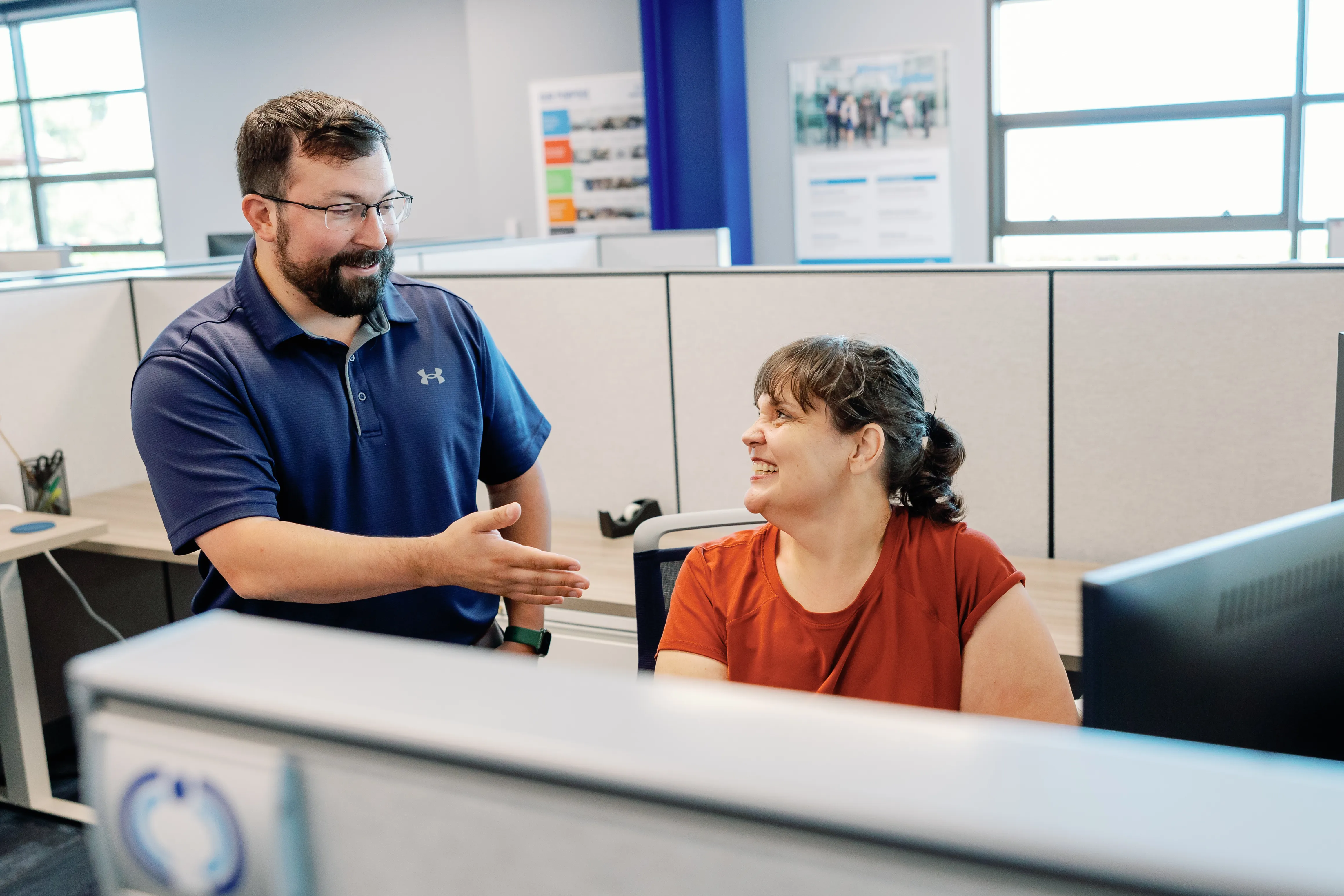 Ein Mann in einem blauen Polohemd begrüßt eine Frau am Schreibtisch in einem modernen Büro. Sie lächelt freundlich.