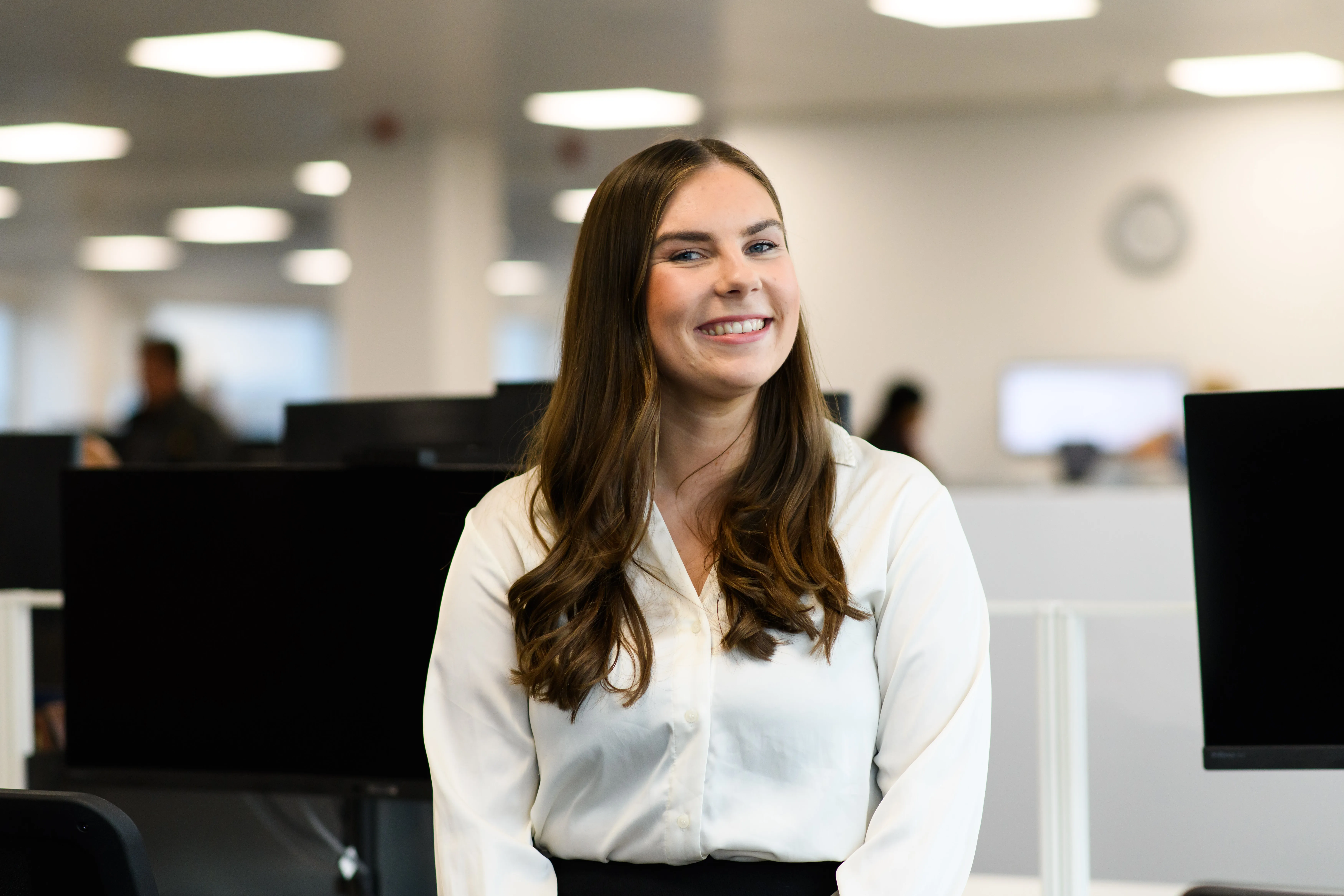 Alt Texa woman with long brown hair, wearing a white blouse, sitting in an office environment. She has a pleasant smile and appears to be in front of computer monitors, with the office space in the background blurred out.t