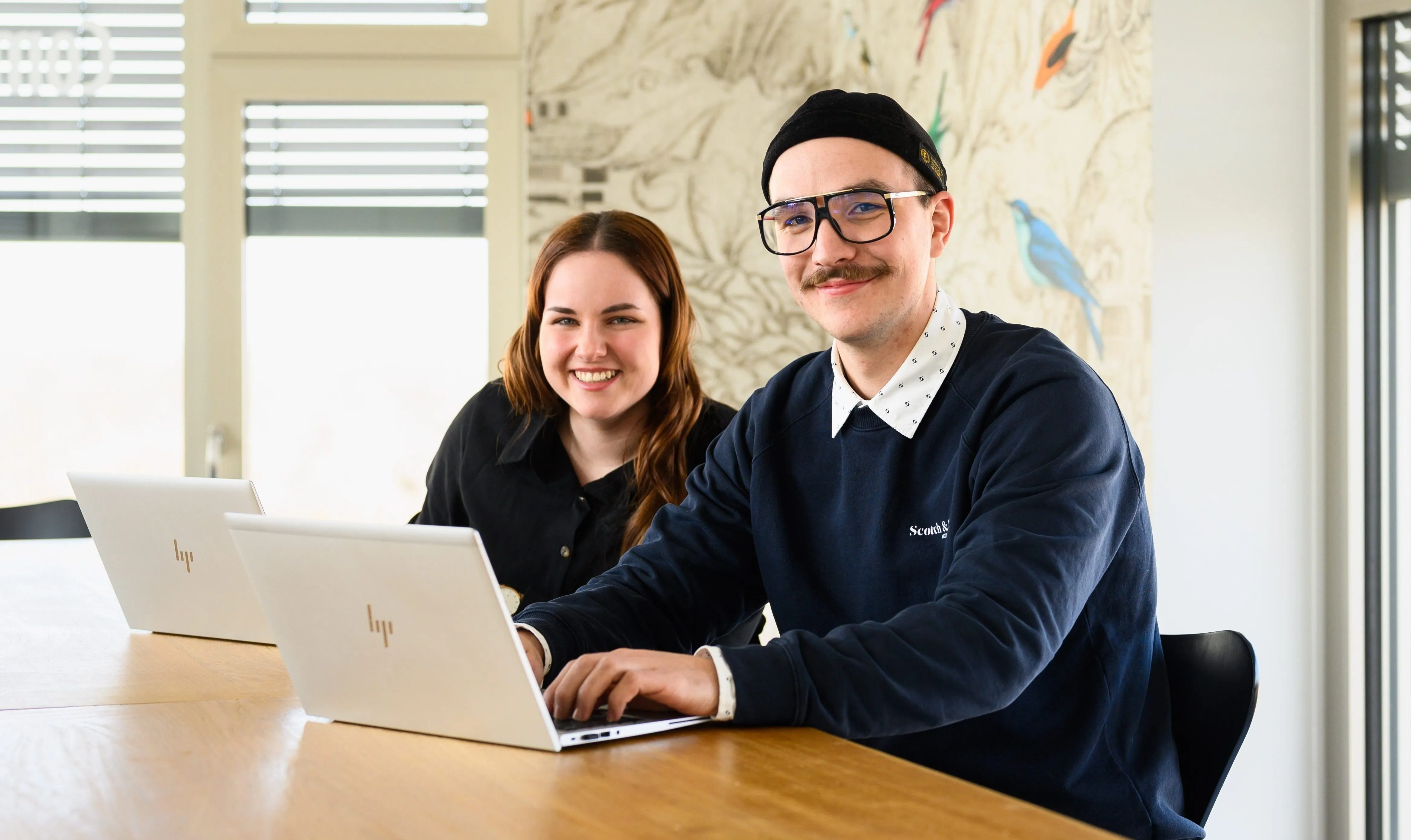 Zwei lächelnde IT-Profis arbeiten gemeinsam an Laptops in einem modernen Büro.