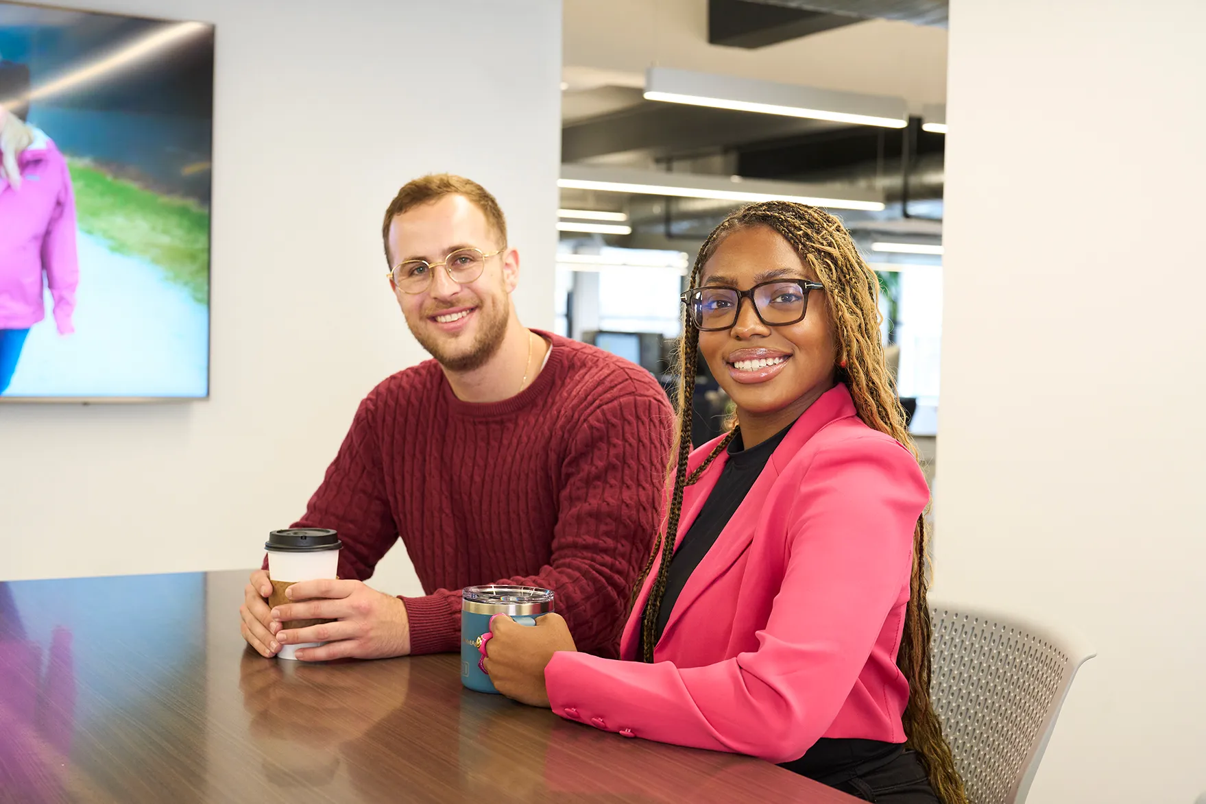 Alt Texa woman with long brown hair, wearing a white blouse, sitting in an office environment. She has a pleasant smile and appears to be in front of computer monitors, with the office space in the background blurred out.t